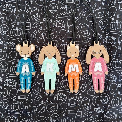 Four Halloween-themed wooden character keychains (a mouse, a cat, a bear, and a rabbit) wearing colorful pajamas with white letters, displayed against a black background with white spooky line drawings.