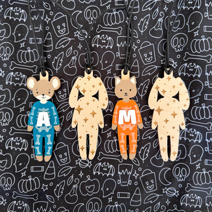 Four wooden bear-shaped pendants with black hanging cords, displayed against a black background patterned with white Halloween icons. Two pendants are dressed in blue and orange pajamas with white letters 'A' and 'M' respectively, while the other two are in natural wood with gold star patterns.