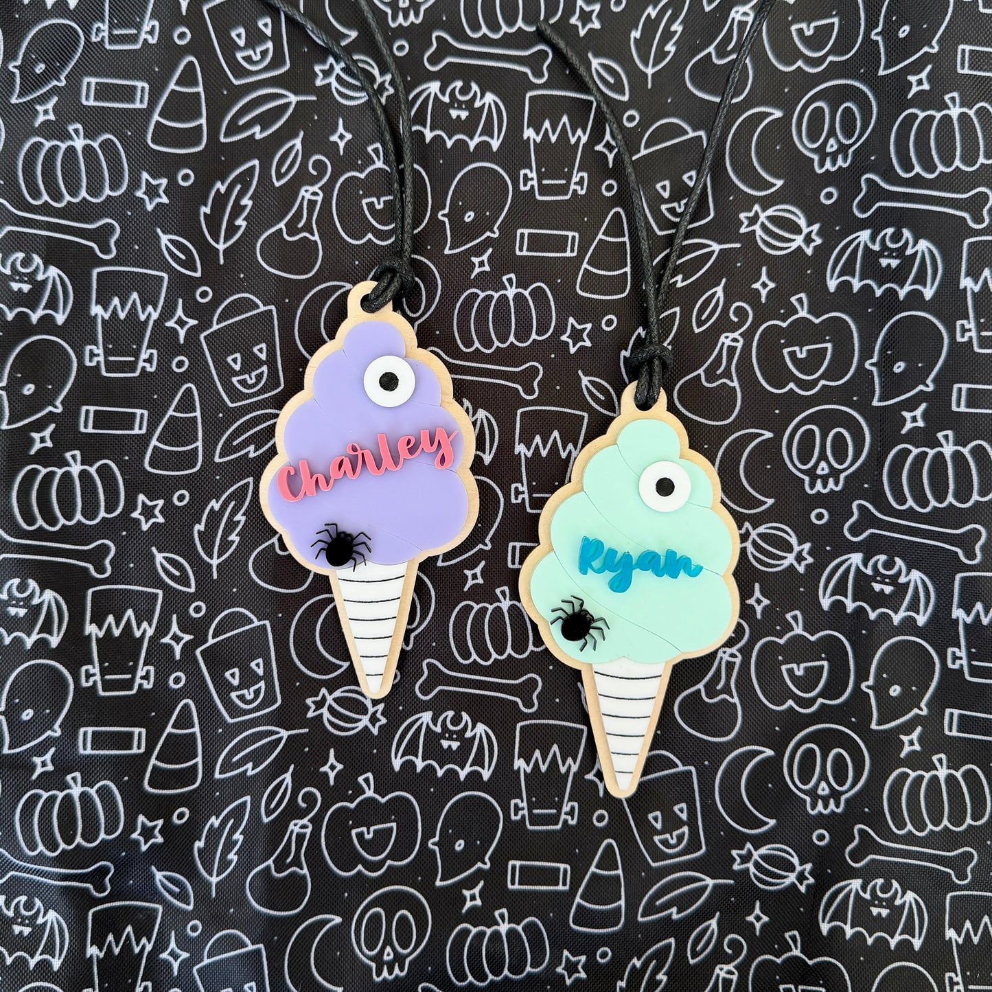 Two personalized ice cream cone charms with Halloween-themed names and spider accents, displayed on a black patterned background.