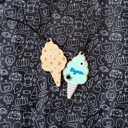 Two Halloween-themed ice cream cone pendants on black cords, displayed on a black background with a white line-art pattern of pumpkins, bats, and ghosts. One cone is tan with gold stars, and the other is mint green with blue text reading 'Ryan' and a black spider detail.